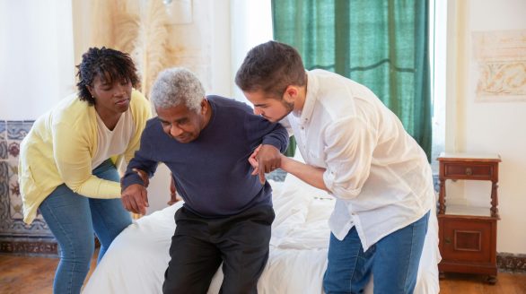An elderly man receiving assistance from caregivers in a cozy home environment.